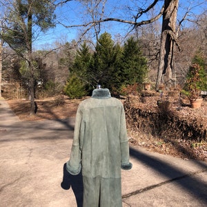May include: A long, green suede coat with a fur collar and cuffs. The coat is on a mannequin and is standing on a paved driveway.