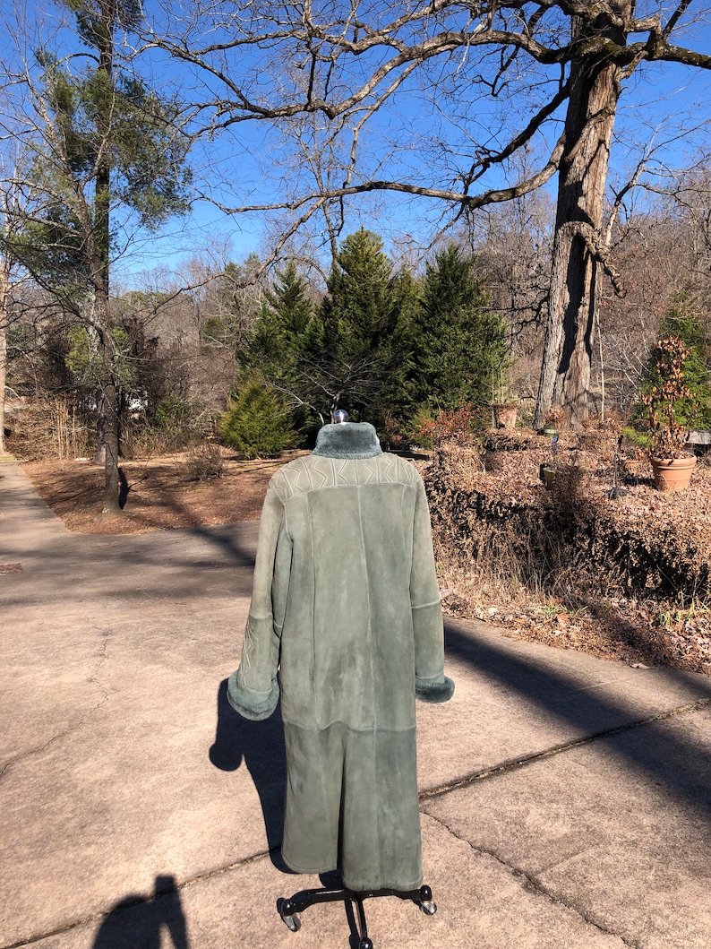May include: A long, green suede coat with a fur collar and cuffs. The coat is on a mannequin and is standing on a paved driveway.