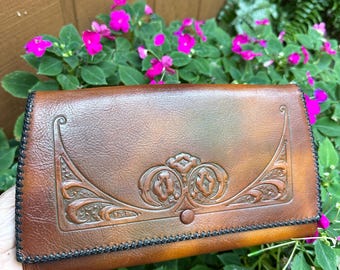 May include: A brown leather wallet with an embossed floral design and black stitching. The wallet is rectangular and has a snap closure. The background features green foliage and pink flowers.