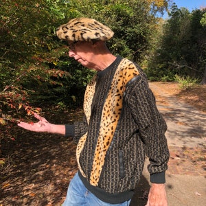 May include: A man wearing a black and brown leopard print sweater with a matching leopard print beret. The sweater has a zipper and a black trim. The man is standing in a wooded area.