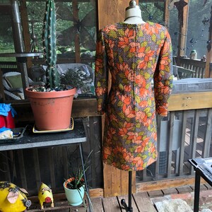 Peut inclure: Une robe vintage &agrave; motif floral brun, vert et orange. La robe a des manches longues et une silhouette ajust&eacute;e. Elle est expos&eacute;e sur un mannequin sur un fond en bois.