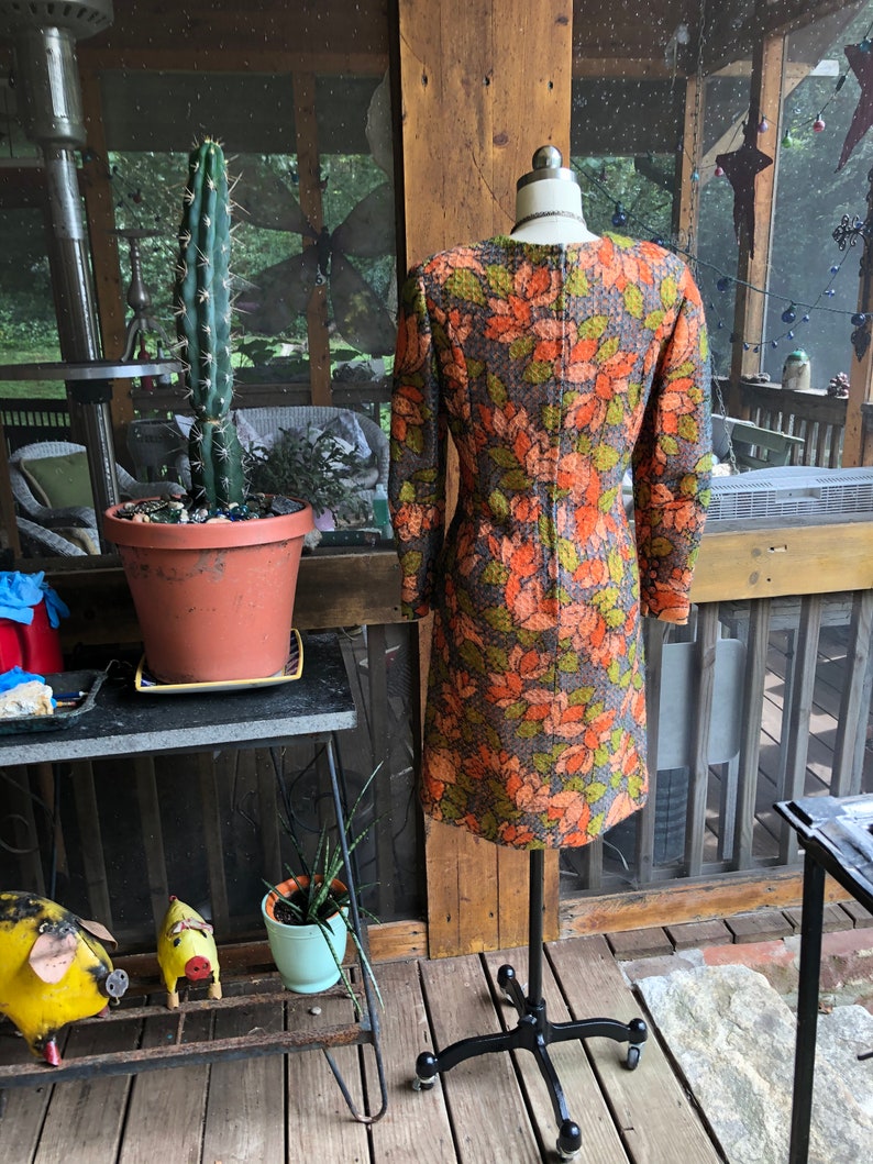Peut inclure: Une robe vintage &agrave; motif floral brun, vert et orange. La robe a des manches longues et une silhouette ajust&eacute;e. Elle est expos&eacute;e sur un mannequin sur un fond en bois.
