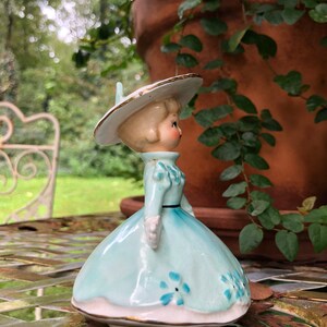 Może przedstawiać: Antyczna figurka porcelanowa przedstawiająca dziewczynkę w niebieskiej sukience z białymi kwiatami i białym kapeluszu z złotą ozdobą. Figurka stoi na stole z metalową powierzchnią.
