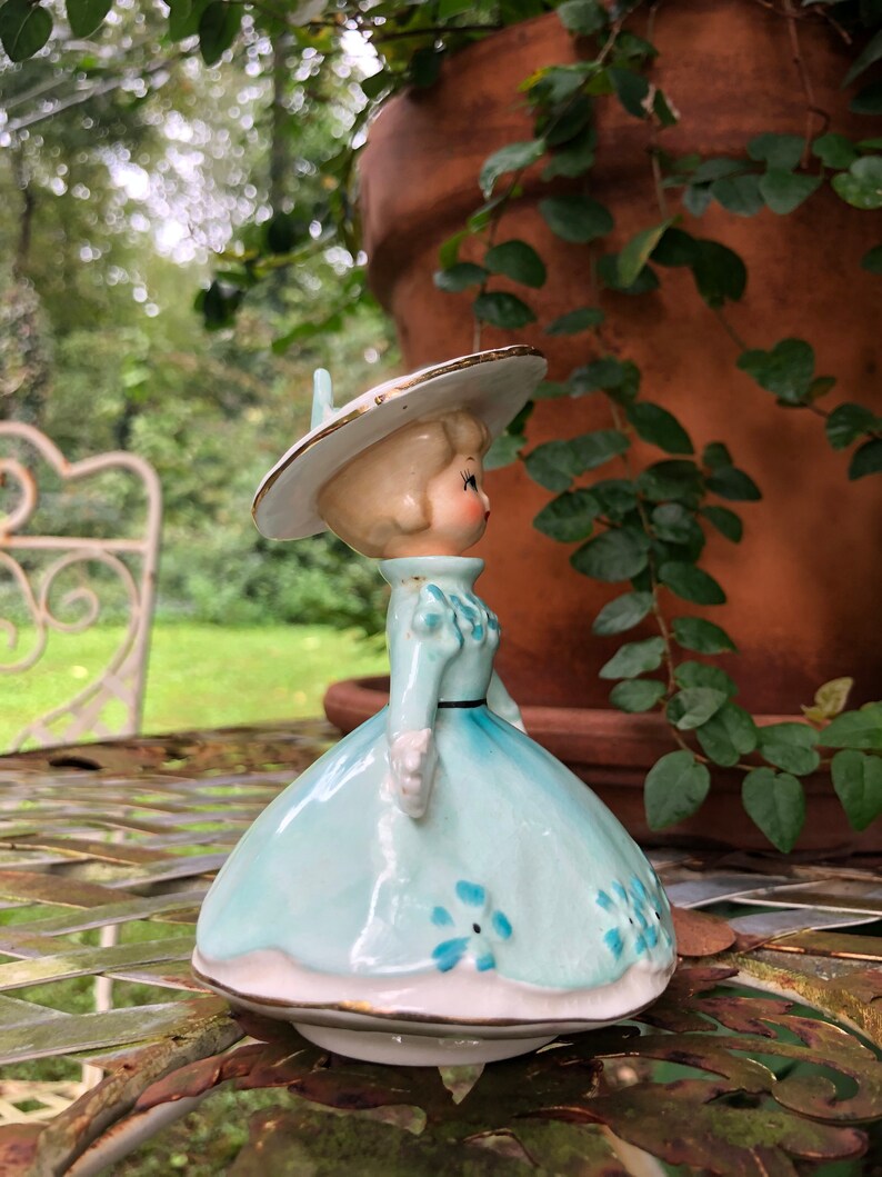 Może przedstawiać: Antyczna figurka porcelanowa przedstawiająca dziewczynkę w niebieskiej sukience z białymi kwiatami i białym kapeluszu z złotą ozdobą. Figurka stoi na stole z metalową powierzchnią.