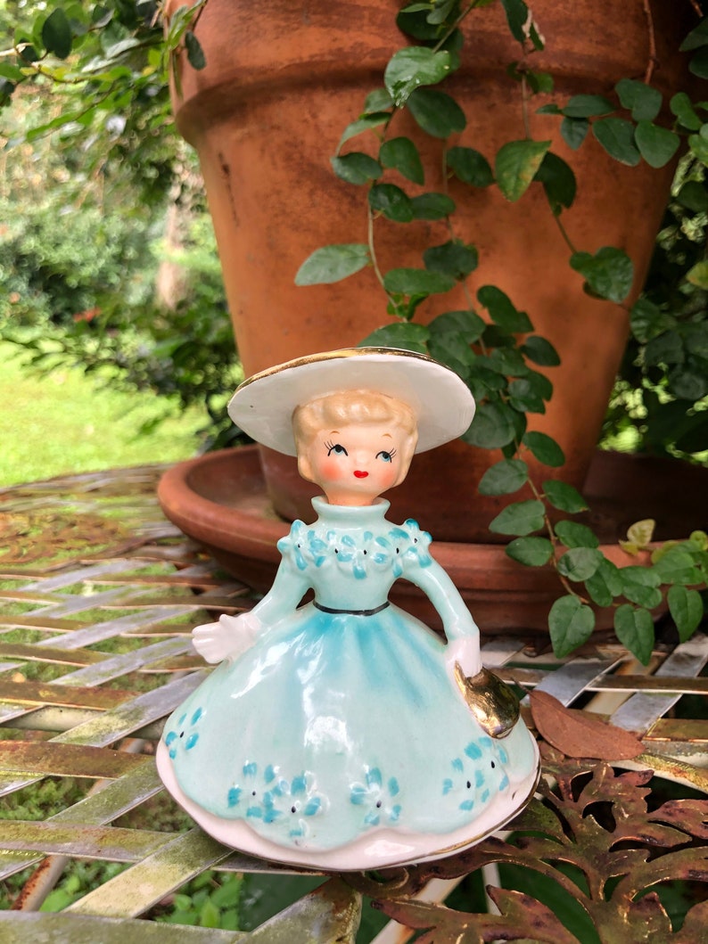 Może przedstawiać: Antyczna figurka porcelanowa przedstawiająca dziewczynkę w jasnoniebieskiej sukience z białymi kwiatami i białym kapeluszu z złotą ozdobą. Trzyma złotą torebkę.
