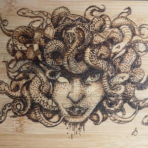 May include: A wood-burned image of Medusa, a mythical Greek figure with snakes for hair, on a wooden cutting board.