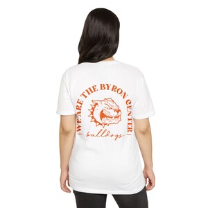May include: White t-shirt with an orange graphic of a bulldog and the text "WE ARE THE BYRON CENTER" in a circular design. Below the graphic, the word "bulldogs" is written in a script font. The shirt is made of a soft material.