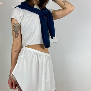 Könnte beinhalten: Ein weißer Knopf-Rock mit abgerundetem Saum. Der Rock ist aus leichtem Stoff und hat eine lockere Passform. Das Model trägt ein weißes Crop-Top und einen marineblauen Pullover.