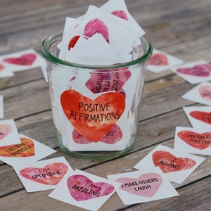 May include: A glass jar filled with small white cards with watercolor hearts and positive affirmations written on them. The jar is labeled "Positive Affirmations". The cards are scattered around the jar on a wooden surface.