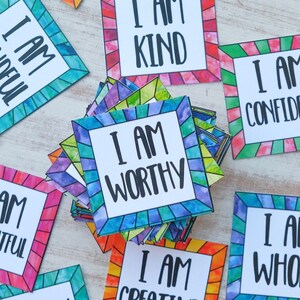 Op de afbeelding: Een stapel kleurrijke affirmatiekaarten met de zin "Ik ben" en een positieve affirmatie op elke kaart. De kaarten hebben een glas-in-loodontwerp met een witte rand.