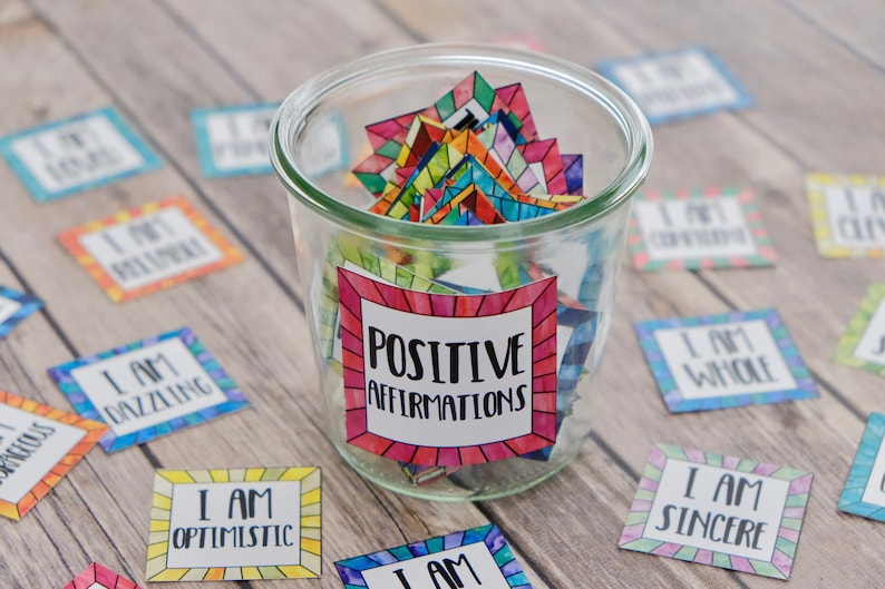Op de afbeelding: Een glazen pot gevuld met kleurrijke vierkante kaartjes met positieve affirmaties erop. De pot heeft een label met de tekst "POSITIVE AFFIRMATIONS".