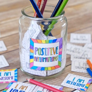 Op de afbeelding: Een glazen pot gevuld met gekleurde papiertjes met positieve affirmaties erop geschreven. De pot is gelabeld met "Positieve Affirmaties" en heeft een regenboogrand. Er zitten verschillende kleurpotloden in de pot. Er liggen ook verschillende affirmatiecards op een houten oppervlak, waaronder "Ik ben geweldig", "Ik ben geliefd", "Ik ben gefocust", "Ik ben veerkrachtig" en "Ik ben fantastisch".
