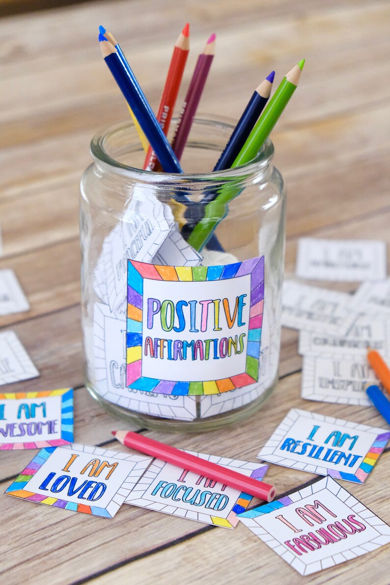 Op de afbeelding: Een glazen pot gevuld met gekleurde papiertjes met positieve affirmaties erop geschreven. De pot is gelabeld met "Positieve Affirmaties" en heeft een regenboogrand. Er zitten verschillende kleurpotloden in de pot. Er liggen ook verschillende affirmatiecards op een houten oppervlak, waaronder "Ik ben geweldig", "Ik ben geliefd", "Ik ben gefocust", "Ik ben veerkrachtig" en "Ik ben fantastisch".