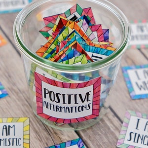 Op de afbeelding: Een glazen pot gevuld met kleurrijke vierkante kaartjes met positieve affirmaties erop. De pot is gelabeld met "POSITIVE AFFIRMATIONS".