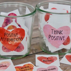 May include: Two glass jars filled with small heart-shaped paper notes. The jars are labeled "Positive Affirmations" and "Think Positive". The notes are decorated with watercolor-like designs in shades of pink and red.