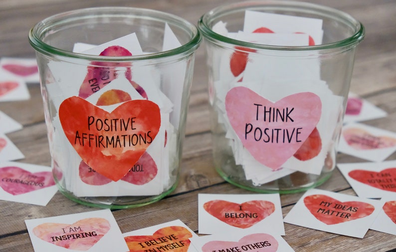 May include: Two glass jars filled with small heart-shaped paper notes. The jars are labeled "Positive Affirmations" and "Think Positive". The notes are decorated with watercolor-like designs in shades of pink and red.