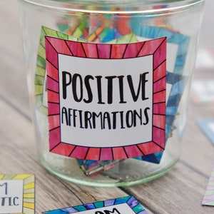 Op de afbeelding: Een glazen pot gevuld met kleurrijke kaartjes waarop positieve affirmaties staan. De pot heeft een label met de tekst "POSITIVE AFFIRMATIONS". Er liggen twee kaartjes buiten de pot, &eacute;&eacute;n zegt "IK BEN GECONCENTREERD" en de andere zegt "IK BEN MAGISCH".