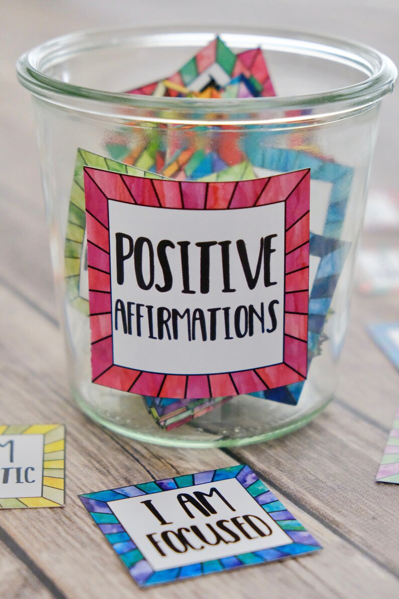 Op de afbeelding: Een glazen pot gevuld met kleurrijke kaartjes waarop positieve affirmaties staan. De pot heeft een label met de tekst "POSITIVE AFFIRMATIONS". Er liggen twee kaartjes buiten de pot, &eacute;&eacute;n zegt "IK BEN GECONCENTREERD" en de andere zegt "IK BEN MAGISCH".