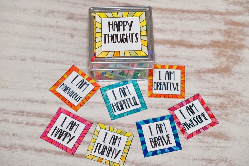 Op de afbeelding: Een doorzichtige plastic doos met een deksel gevuld met kleurrijke papieren vierkanten. De doos heeft een label met de tekst "Happy Thoughts". Rondom de doos liggen acht kleurrijke papieren vierkanten met positieve affirmaties erop, zoals "Ik ben creatief" en "Ik ben hoopvol".