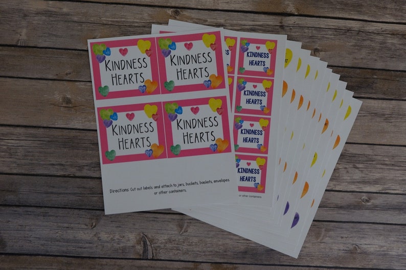 Pu&ograve; includere: Un foglio di etichette stampabili con sfondo rosa e cuori colorati che circondano il testo "Kindness Hearts". Le etichette sono progettate per essere ritagliate e attaccate a barattoli, secchi, cesti, buste o altri contenitori.
