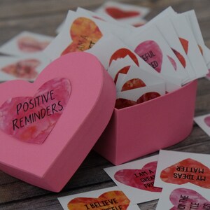 May include: A pink heart-shaped box with the words "Positive Reminders" on the lid. The box is filled with small, heart-shaped cards with positive affirmations written on them.