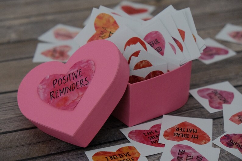 May include: A pink heart-shaped box with the words "Positive Reminders" on the lid. The box is filled with small, heart-shaped cards with positive affirmations written on them.