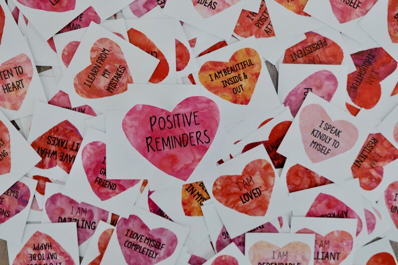 May include: A collection of pink and red watercolor heart-shaped cards with positive affirmations printed on them. The cards include phrases like "I am loved", "I speak kindly to myself", and "I love myself completely".