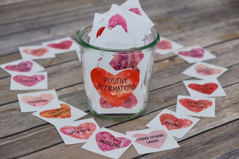 May include: A glass jar filled with small white cards with watercolor hearts and positive affirmations written on them. The jar is labeled "Positive Affirmations". The cards are scattered around the jar on a wooden surface.