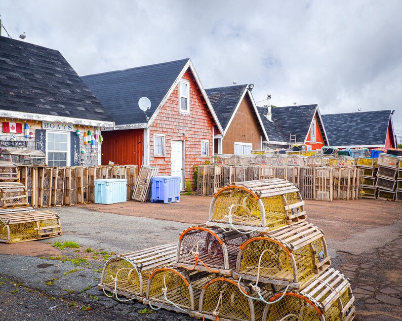May include: A row of colorful houses with wooden lobster traps stacked in front of them. The houses have black roofs and white trim. The lobster traps are made of wood and wire mesh.
