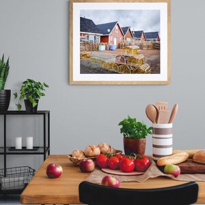 May include: A framed photograph of a row of red houses with a pile of lobster traps in the foreground. The houses are in a coastal town and the traps are stacked high.