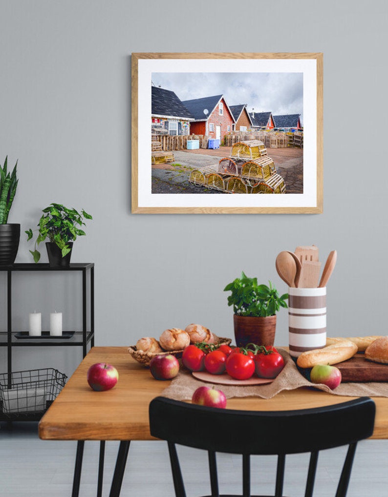 May include: A framed photograph of a row of red houses with a pile of lobster traps in the foreground. The houses are in a coastal town and the traps are stacked high.