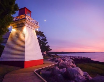 May include: A white lighthouse with a red roof stands on a rocky shore at sunset. The sky is a vibrant pink and purple, and the water is calm and still.