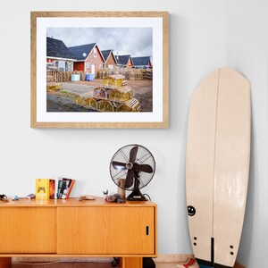 May include: A framed photograph of a row of red houses with a pile of lobster traps in the foreground. The houses are in a coastal town and the sky is cloudy.