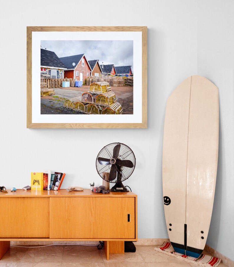 May include: A framed photograph of a row of red houses with a pile of lobster traps in the foreground. The houses are in a coastal town and the sky is cloudy.