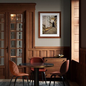 May include: A dining room with a wooden table and chairs. The table has a dark wood top and a black metal base. The chairs are upholstered in a dark red fabric. A framed photograph of a cobblestone street hangs on the wall. The room has a dark wood floor and a light gray wall.