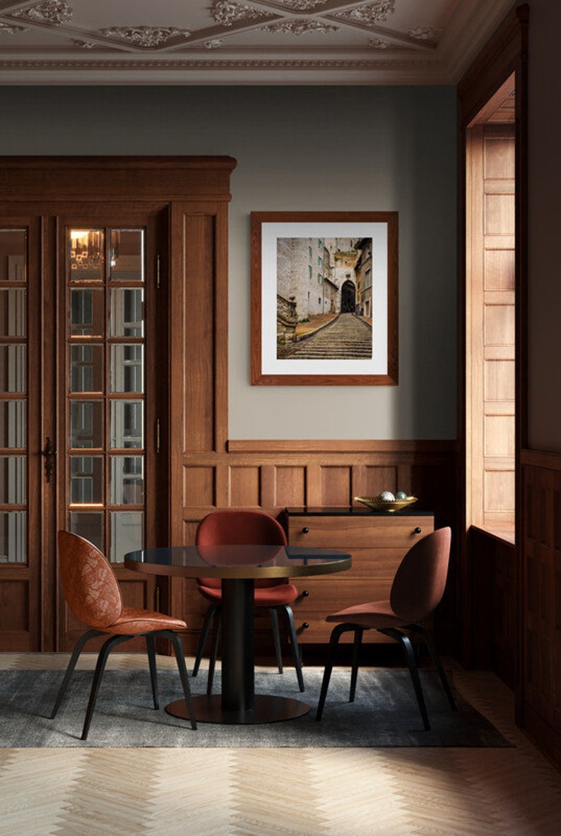 May include: A dining room with a wooden table and chairs. The table has a dark wood top and a black metal base. The chairs are upholstered in a dark red fabric. A framed photograph of a cobblestone street hangs on the wall. The room has a dark wood floor and a light gray wall.