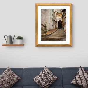 May include: A framed photograph of a narrow, cobblestone street in a European city. The street leads up a set of stone steps to an archway. The buildings on either side of the street are made of stone and have windows with green shutters.