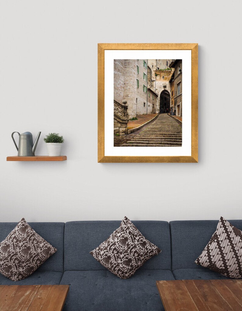 May include: A framed photograph of a narrow, cobblestone street in a European city. The street leads up a set of stone steps to an archway. The buildings on either side of the street are made of stone and have windows with green shutters.