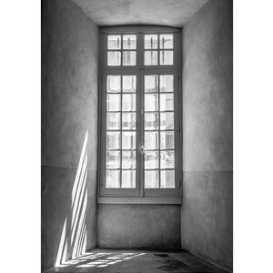 Op de afbeelding: Een zwart-witfoto van een raam met uitzicht op een gebouw buiten. Het raam bevindt zich in een kamer met witte muren en een betonnen vloer. De zon schijnt door het raam en creëert lichtstrepen op de muur.