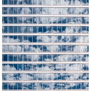 May include: A high-rise building with many windows reflecting a blue sky with white clouds.