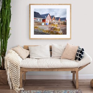 May include: A framed photograph of a row of red houses with a pile of lobster traps in the foreground. The houses are in a coastal town with a cloudy sky.