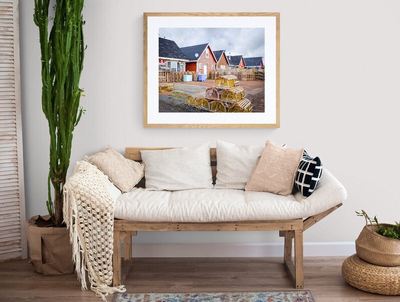 May include: A framed photograph of a row of red houses with a pile of lobster traps in the foreground. The houses are in a coastal town with a cloudy sky.