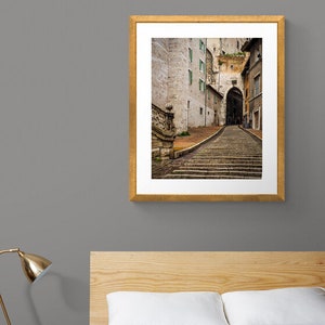May include: A framed photograph of a narrow, cobblestone street in a European city. The street leads up a set of stone steps to an arched doorway. The buildings on either side of the street are old and have a weathered, rustic appearance.