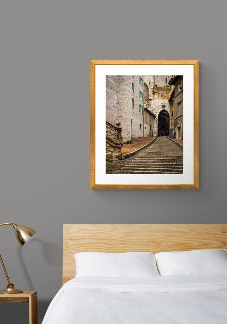 May include: A framed photograph of a narrow, cobblestone street in a European city. The street leads up a set of stone steps to an arched doorway. The buildings on either side of the street are old and have a weathered, rustic appearance.