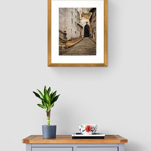 May include: A framed photograph of a narrow, cobblestone street in a European city. The street is lined with old buildings and leads to an archway. The photograph is in a gold frame.