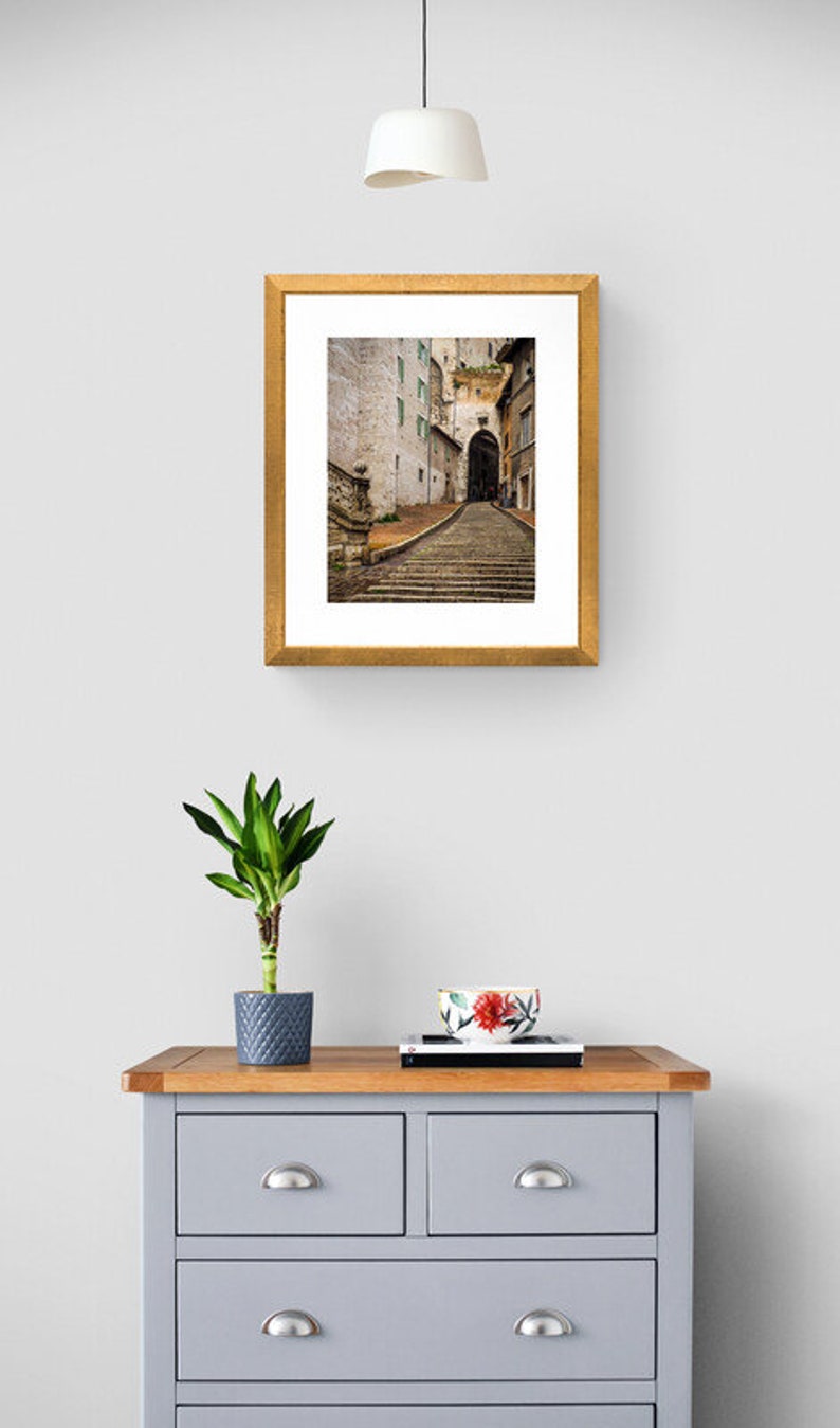 May include: A framed photograph of a narrow, cobblestone street in a European city. The street is lined with old buildings and leads to an archway. The photograph is in a gold frame.