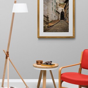 May include: A red armchair with wooden legs and a white side table with a wooden top. A gold framed picture of a cobblestone street with a stone archway hangs on a gray wall. A wooden floor lamp with a white shade and a leather strap is in the corner.