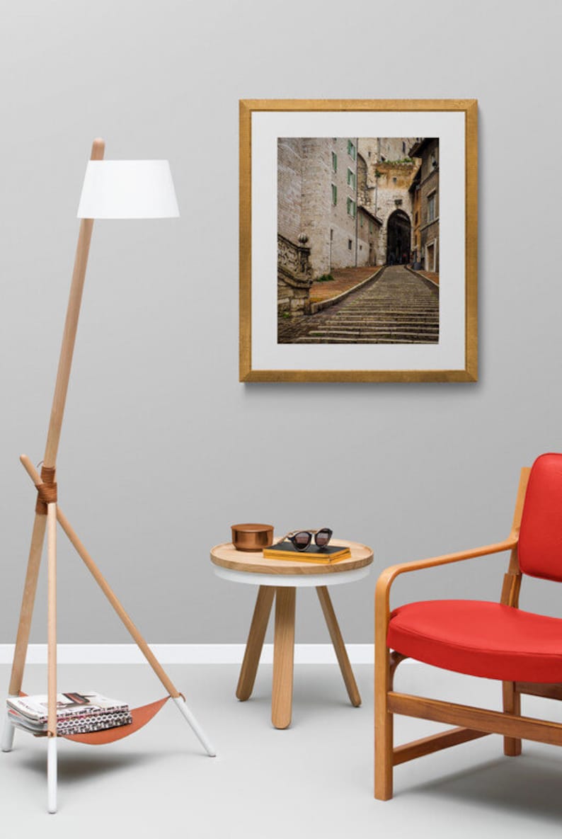 May include: A red armchair with wooden legs and a white side table with a wooden top. A gold framed picture of a cobblestone street with a stone archway hangs on a gray wall. A wooden floor lamp with a white shade and a leather strap is in the corner.