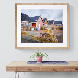 May include: A photograph of a row of red houses with a pile of lobster traps in the foreground. The houses are all painted red and have white trim. The lobster traps are stacked in a pyramid shape and are made of wood and rope.
