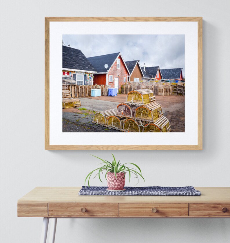 May include: A photograph of a row of red houses with a pile of lobster traps in the foreground. The houses are all painted red and have white trim. The lobster traps are stacked in a pyramid shape and are made of wood and rope.